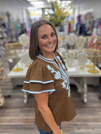 Bluebell bloom embroidered top -  features light blue floral embroidery on the front, matching blue trim along the round neckline and tiered flutter sleeves, and a split neckline