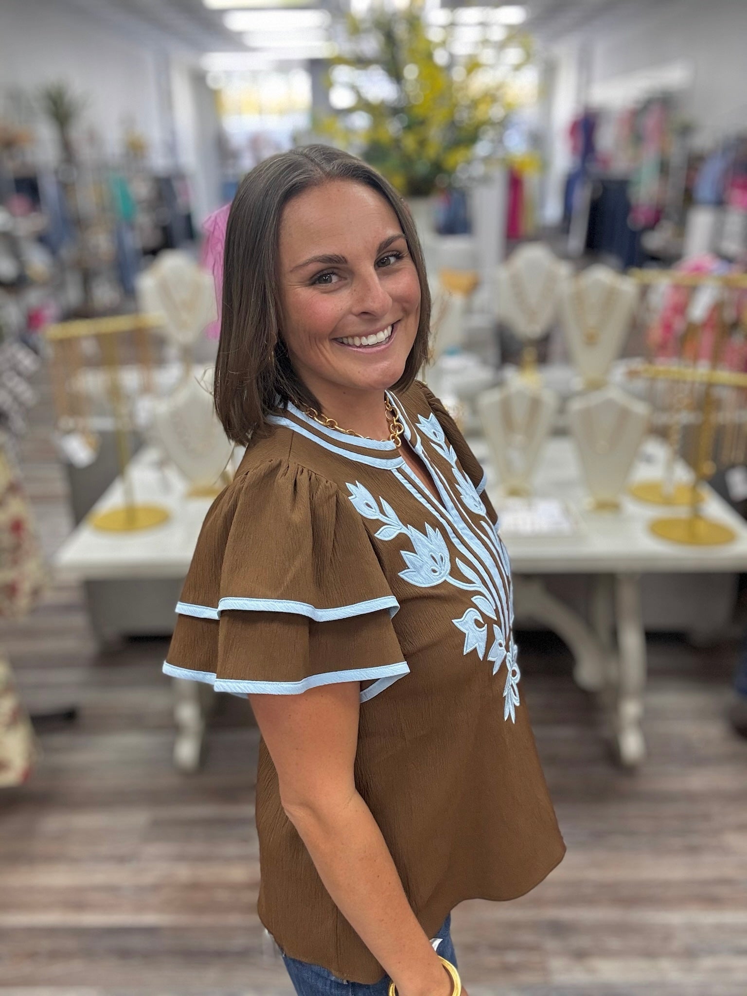 Bluebell bloom embroidered top -  features light blue floral embroidery on the front, matching blue trim along the round neckline and tiered flutter sleeves, and a split neckline