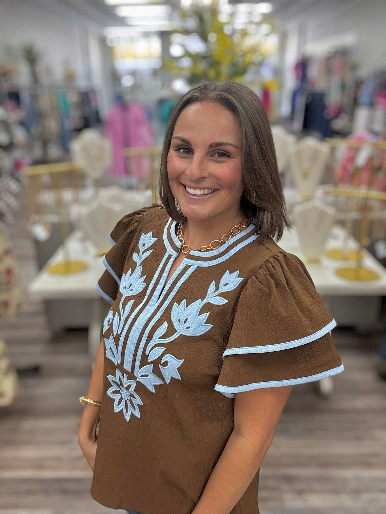 Bluebell bloom embroidered top -  features light blue floral embroidery on the front, matching blue trim along the round neckline and tiered flutter sleeves, and a split neckline